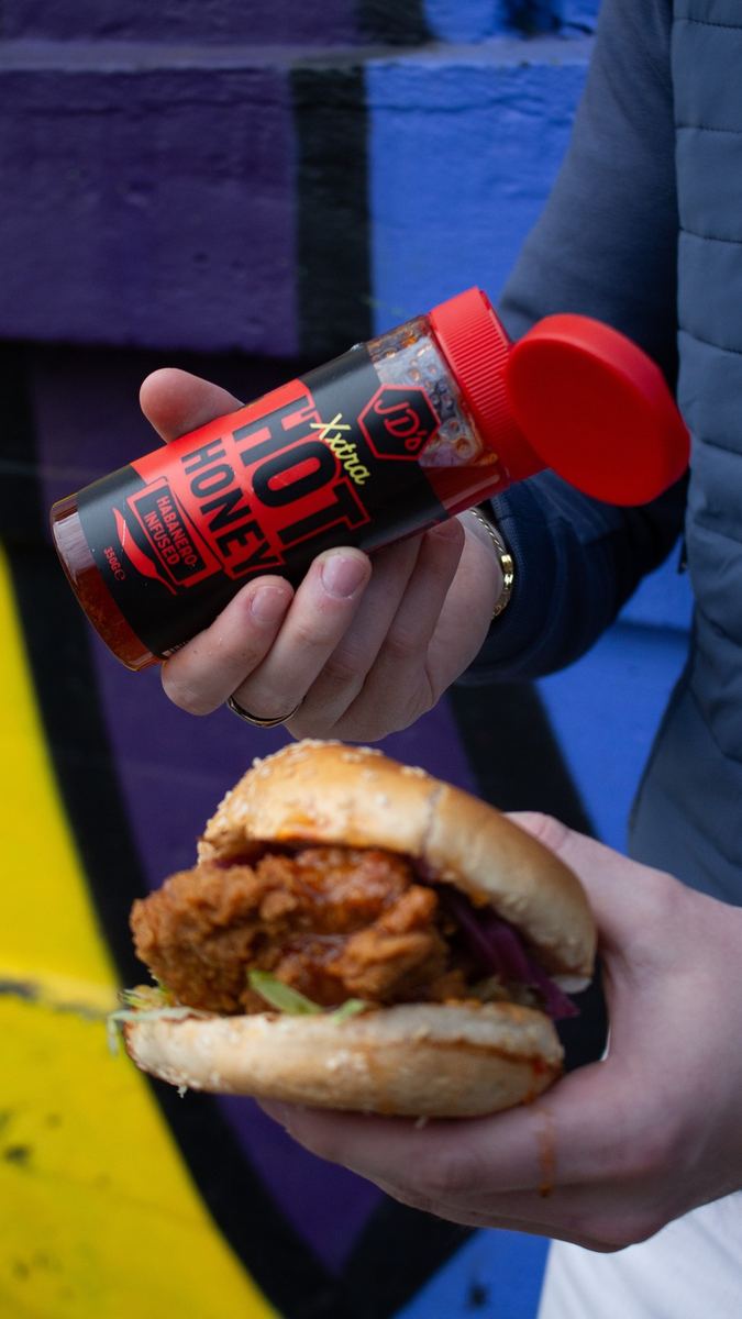 Hand holding a JD's hot honey bottle beside a fried chicken burger